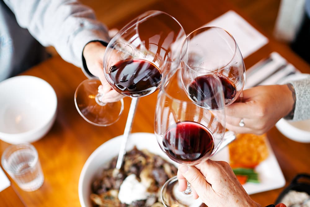 Three glasses of red wine are clinking in celebration over a table with food.