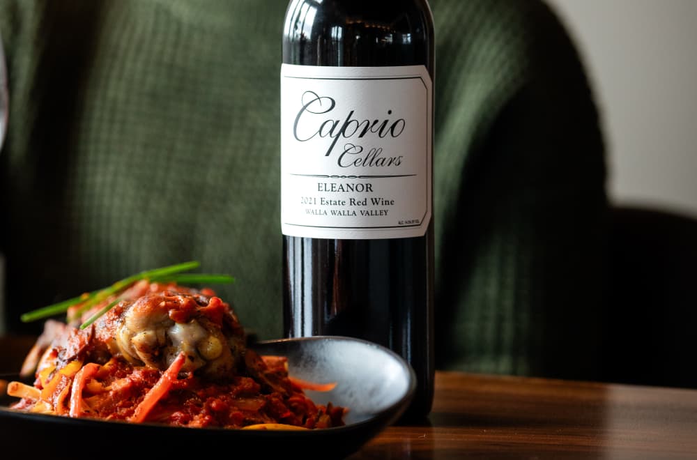 A bottle of Caprio Cellars Eleanor 2021 Estate Red Wine beside a plate of pasta with sauce and vegetables.
