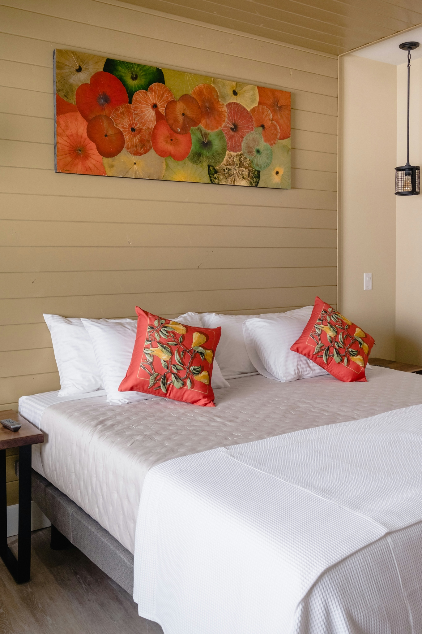 A close-up view of the Brix Suite king-sized bed which can be reconfigured into two twin beds showing four white pillows, a dark beige quilted coverlet and a white waffle throw. A live-edge black walnut bedside table on the side with simple black metal legs, bedside mood lights in a black cage metal finish, original artwork done by your hostess, Naina, on dried lotus leaves, in shades of orange, green and gold, cushions with golden pears and green leaves that pick up the artwork colors, dark cream walls, a light brown wood canopy, The floor is light gray laminate plank.