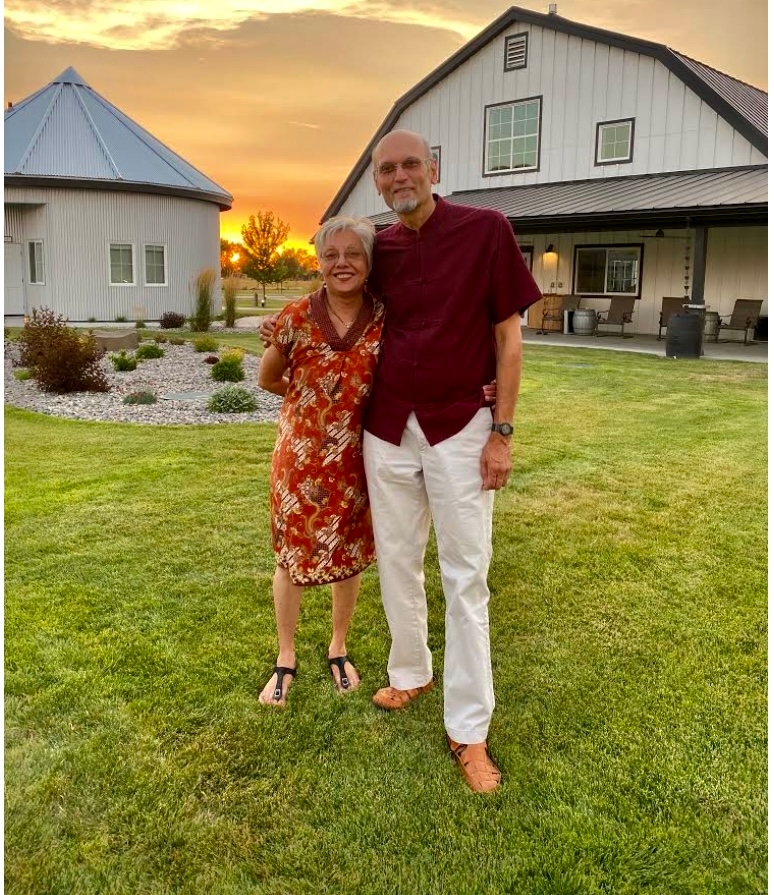 A man and woman standing on the lawn outside of a large white barn home at sunset