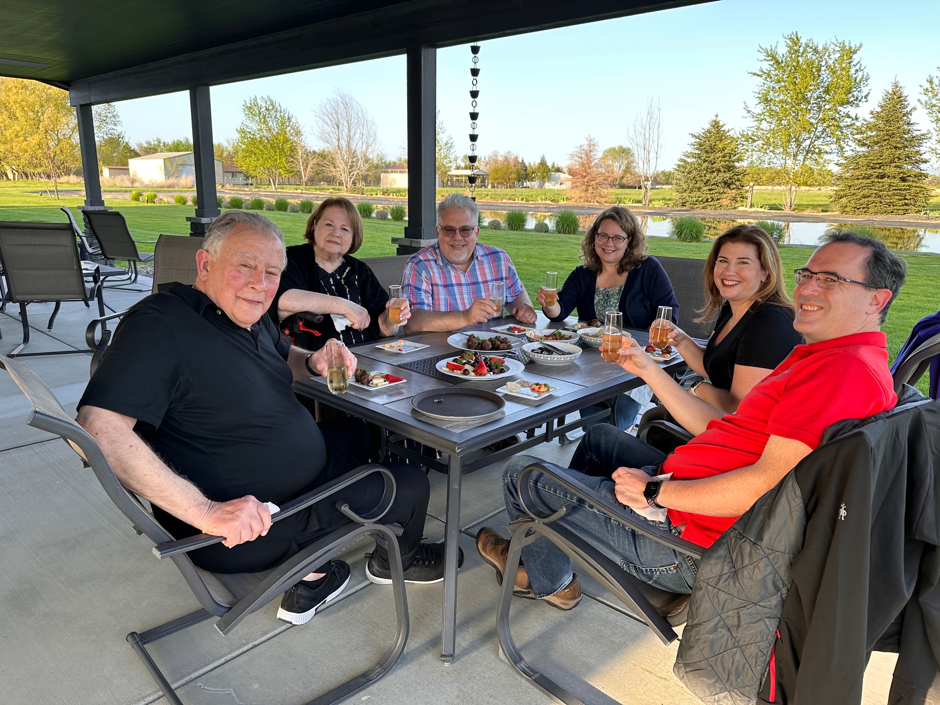 A group of people enjoying a meal at an outdoor patio table next to a large lawn and pond