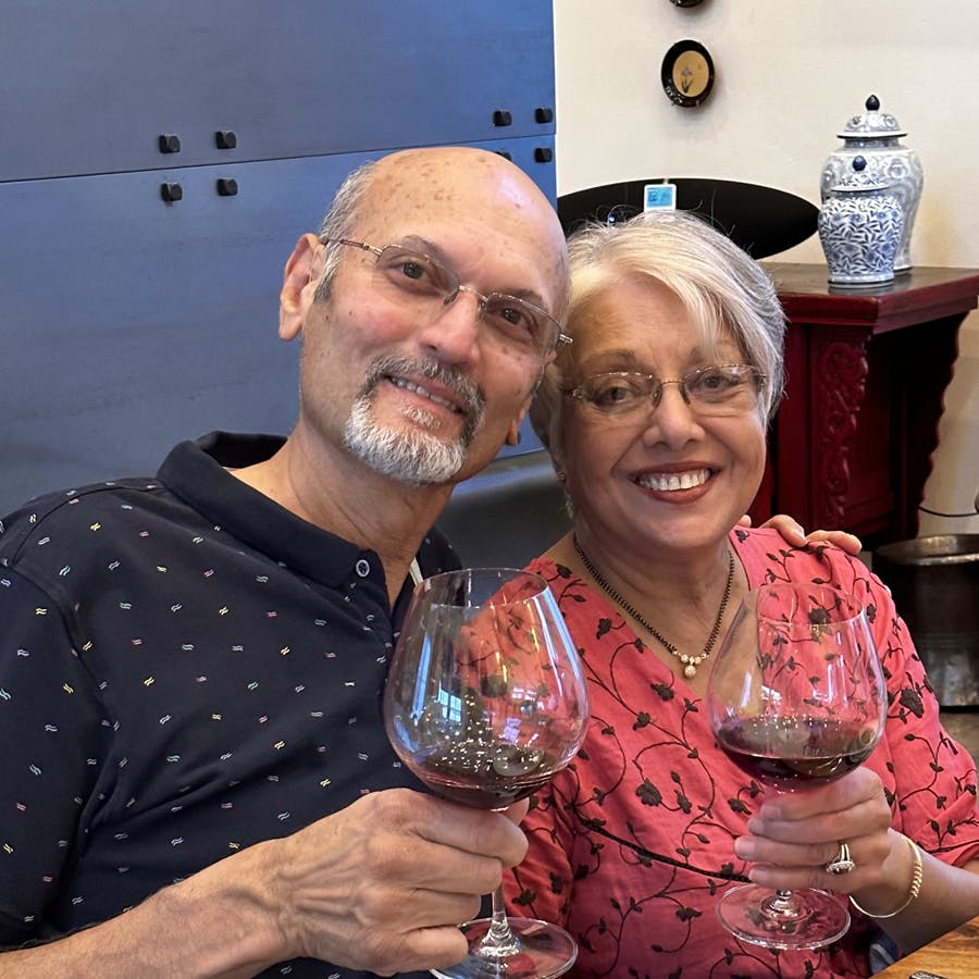 Innkeepers Anand in a blue shirt, and Naina in a peach shirt hold stemmed wine glasses containing red wine.