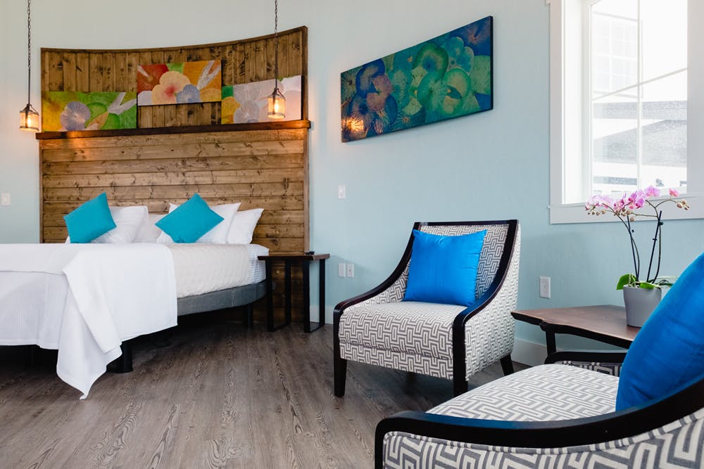 Bed with Dark wood headboard, white linens, teal pillows, geometric grey and white cushioned chairs with dark wood trim and colorful artwork on wall