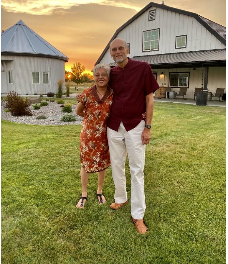 A man and woman standing on the lawn outside of a large white barn home at sunset