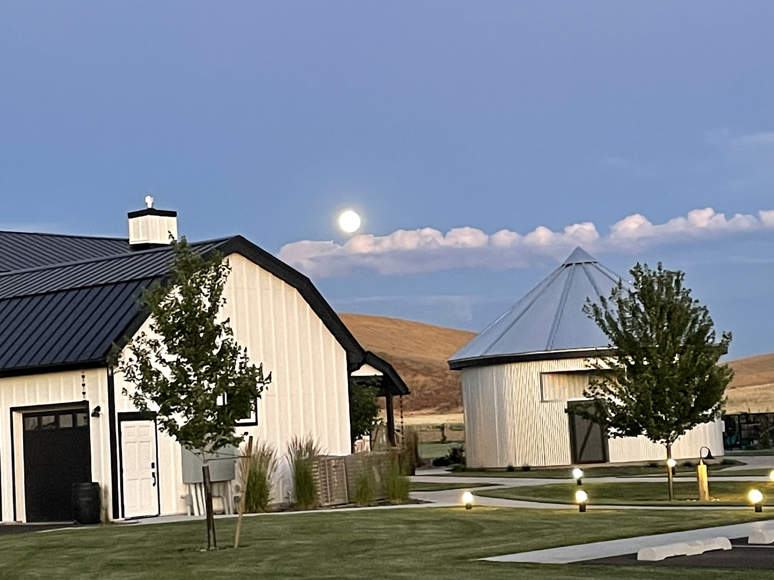 A moon glowing in a bright blue sky over a white barn and silo with grass and sidewalks