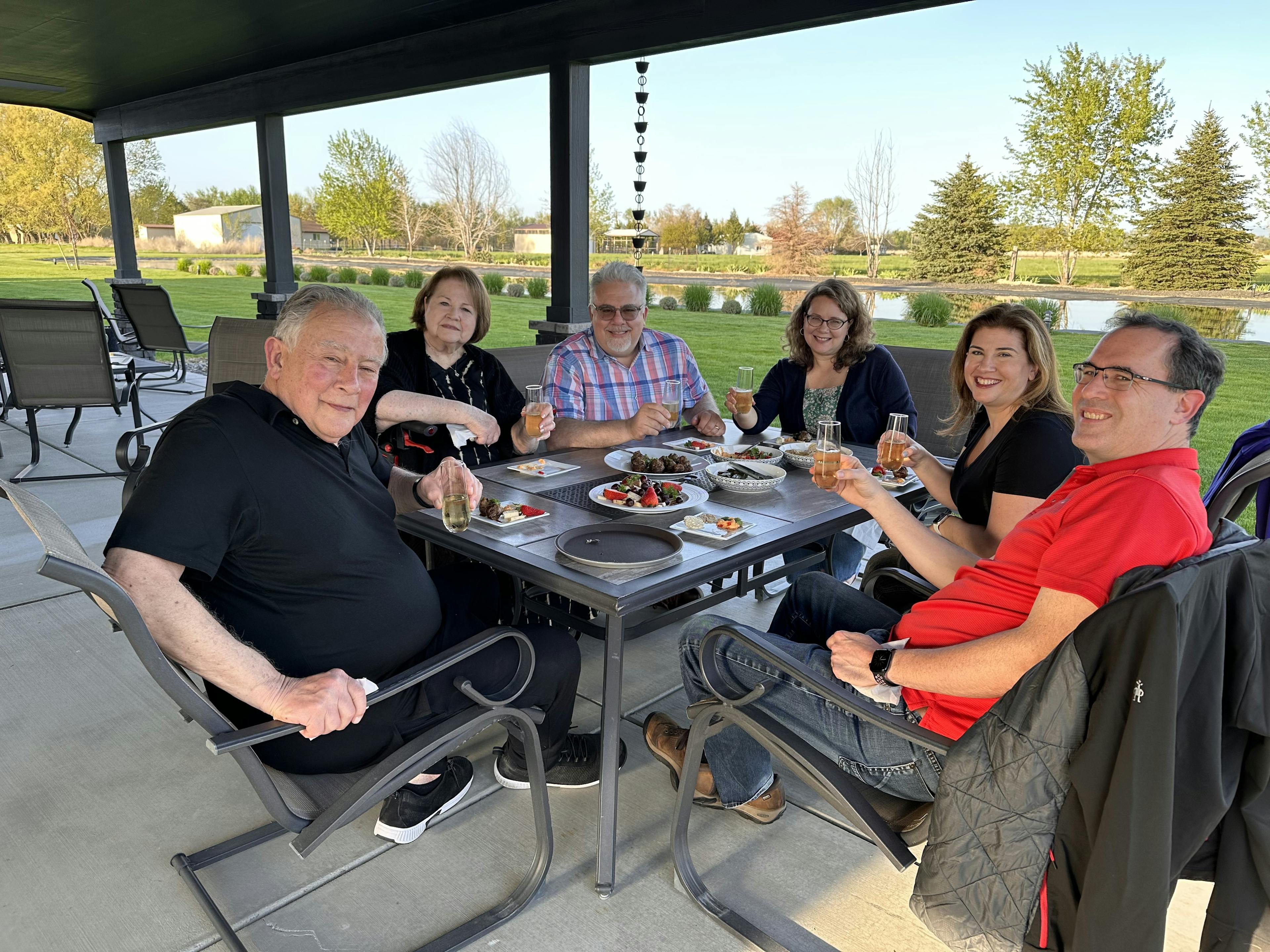 A group of people enjoying a meal at an outdoor patio table next to a large lawn and pond