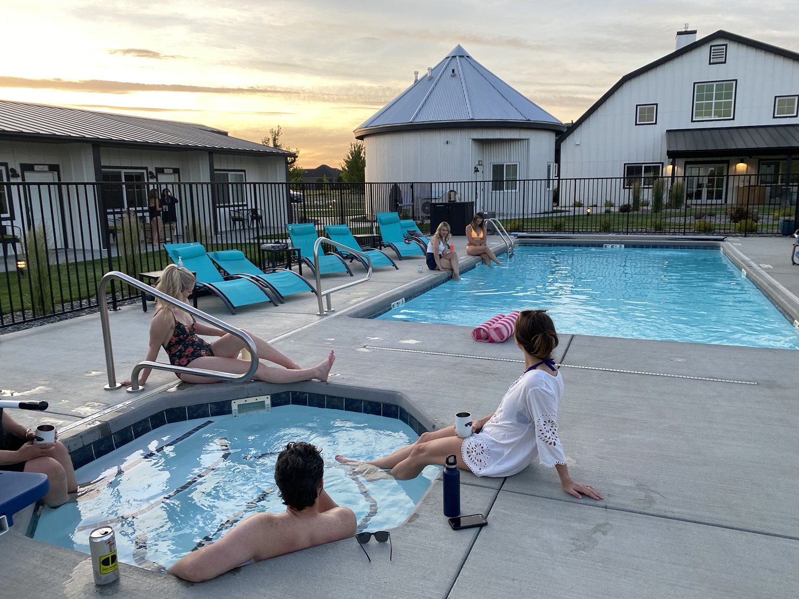 Six people lounging by a outdoor pool and hot tub next to a large white barn home