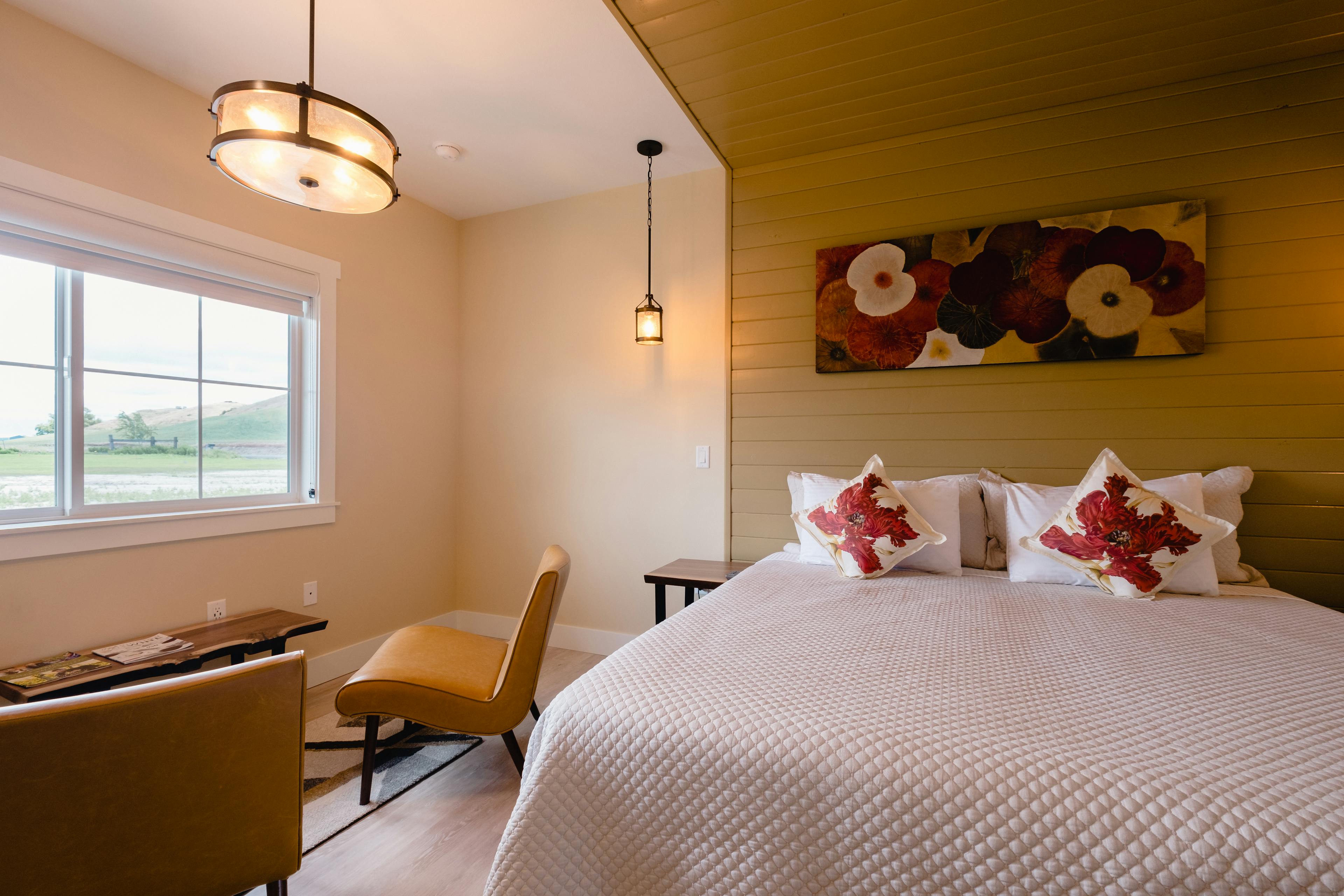 A view of the Tannin Suite showing a King bed which can be reconfigured into twin beds, a live-edge black walnut bedside table with simple black metal legs, comfortable seats in a tan leather, mood lights in an antique glass, wood and black metal finish, original artwork done by your hostess, Naina, on dried lotus leaves, in shades of red, cream, brown and charcoal, cushions printed with a large red flower and brown leaves that pick up the artwork colors, cream painted walls, light brown wood canopy, a white ceiling fan and a white barn door hanging on black metal hardware, leading to the bathroom. The floor is light gray laminate plank. This view shows the wall-mounted 43" flat screen Roku-enabled TV, the mini-split room air conditioner for total control of the temperature. It also shows a wine barrel with a cut-out to fit a small wine fridge. On top of the barrel are two stemless logo wine glasses and wine openers. Beyond the barrel is a live-edge black walnut desk which carries the Nespresso coffee machine and tea kettle with a tea bag selection. The overall look is one of uncluttered elegance and a harmonized color palette.