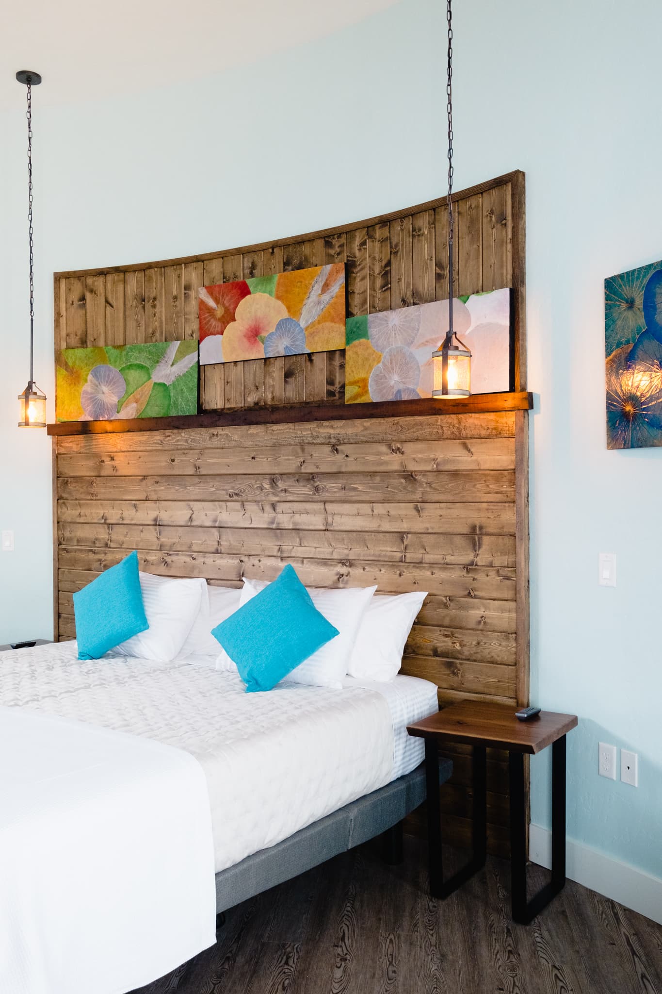 A close-up view of The Granary king-sized bed which can be reconfigured into two twin beds showing four white pillows, a white waffle coverlet. A live-edge black walnut bedside table on the side with simple black metal legs, bedside mood lights in antique wood and metal finish, two works of original art done by your hostess, Naina, on dried lotus leaves, one in shades of blue, turquoise and green and the other depicting the four seasons done in shades of green, beige, fall colors transitioning to silver and snow white. The bed cushions are marine blue and green and white - they pick up the artwork colors, pale blue walls, a natural wood headboard. The floor is light gray laminate plank.