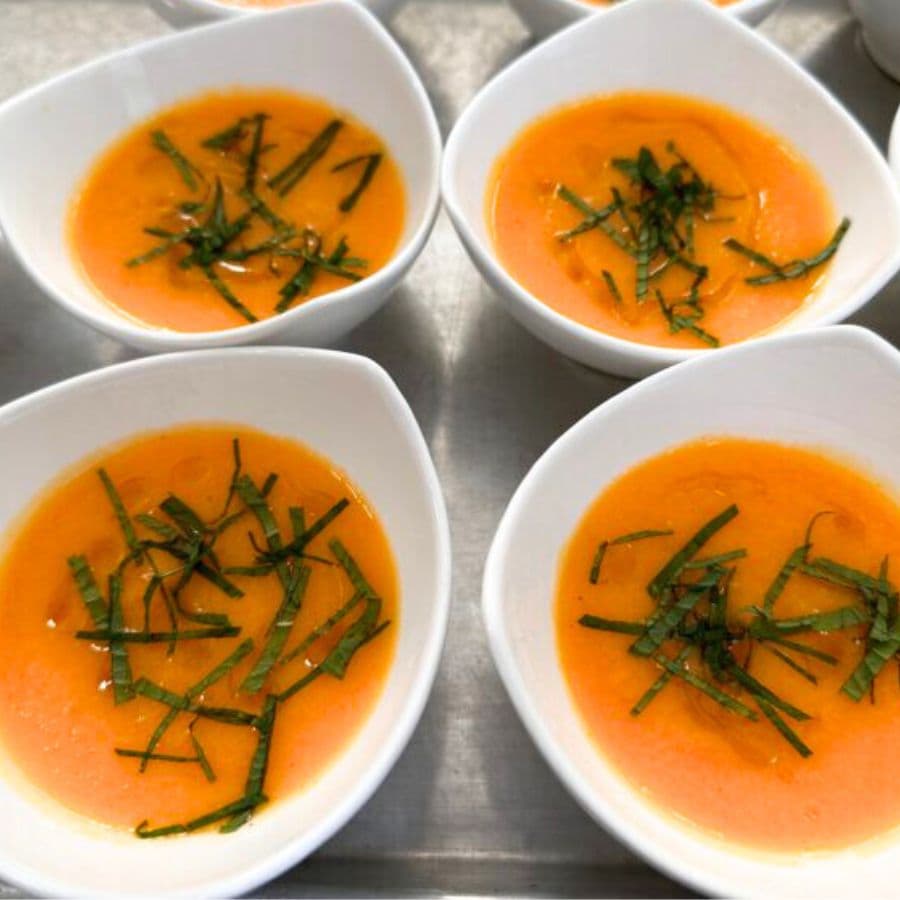 Four white bowls with a bright orange cantaloupe soup and spearmint leaves