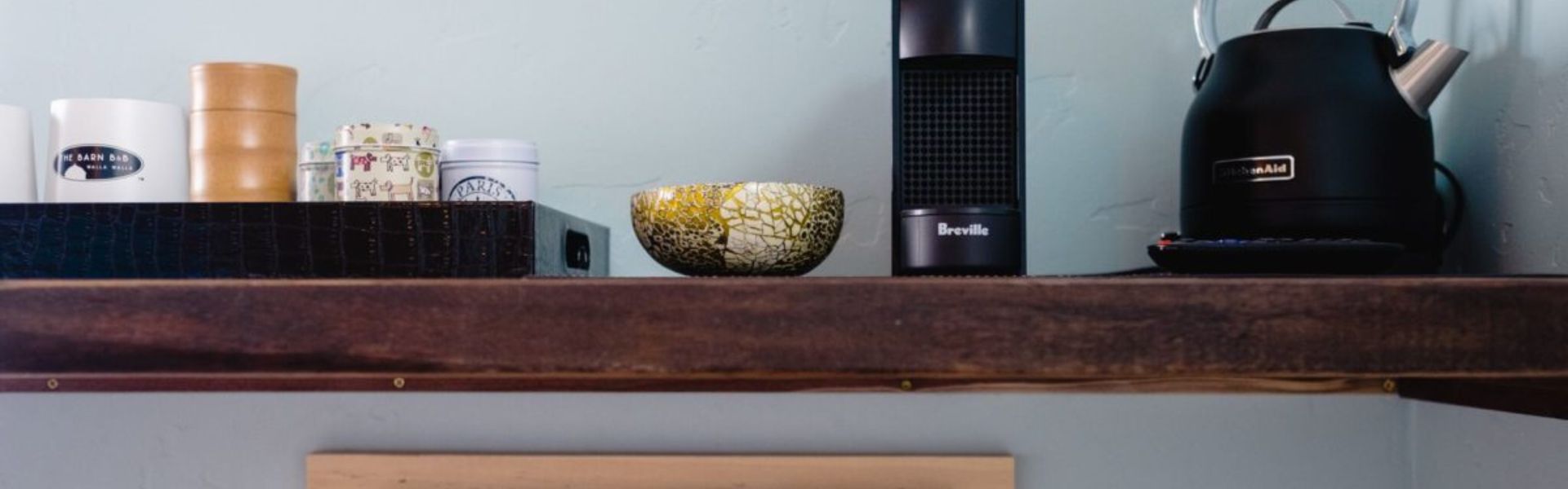 A wooden floating shelf with a tray of coffee mugs and tea canisters, tea kettle and coffee maker