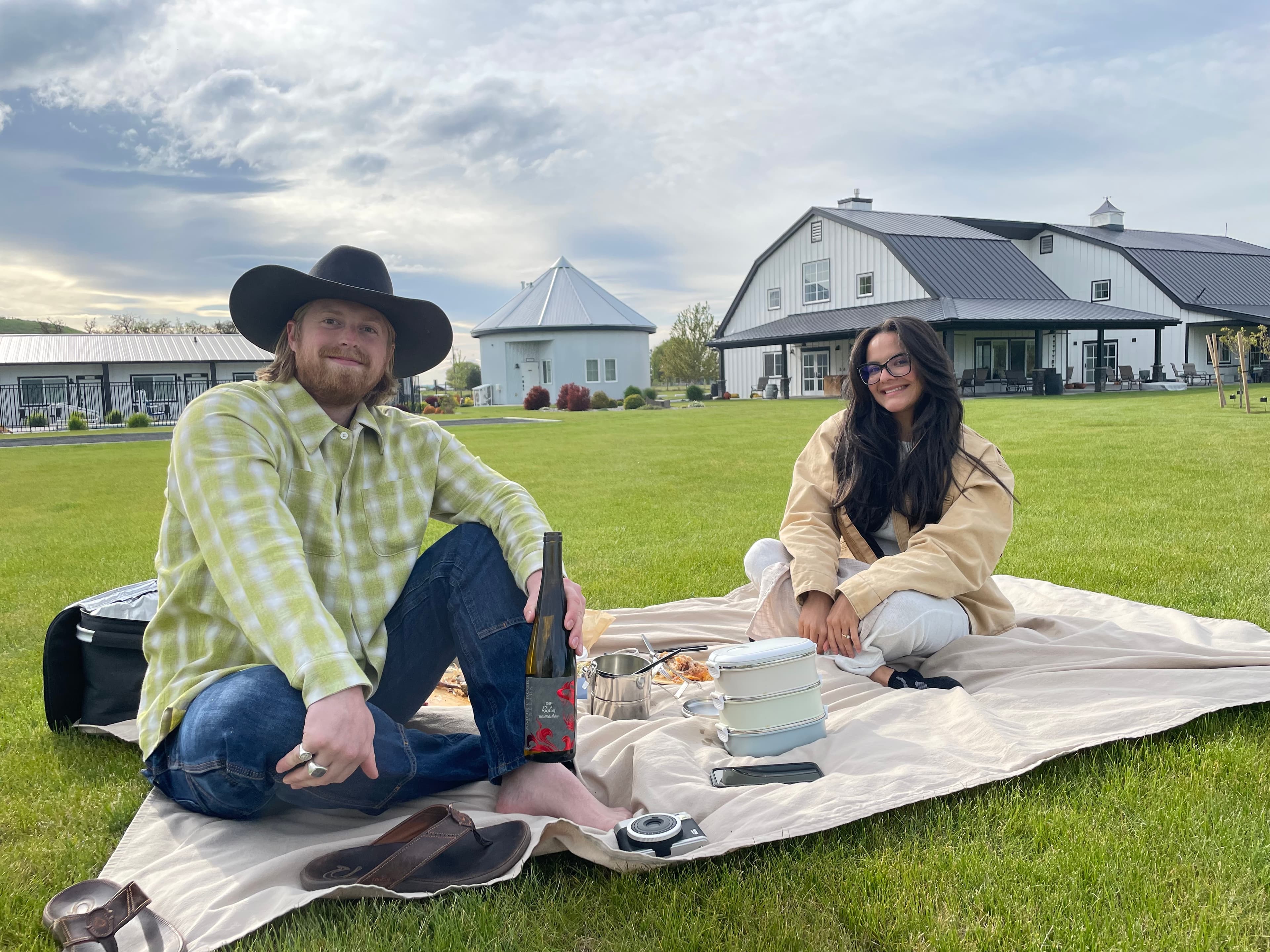 A woman and a man in a cowboy hat sitting on a blanket outdoors enjoying a picnic