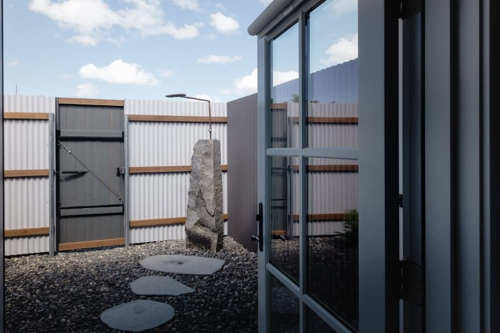 Outdoor shower, gravel and stepping stones on ground, enclosed by aluminum color siding and wood fence.