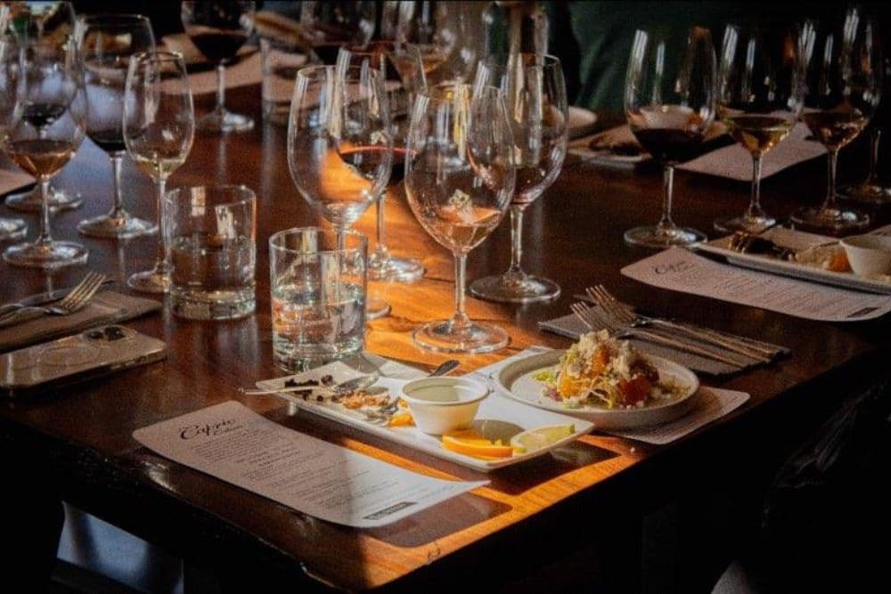 Dining table at a restaurant filled with wine glasses and small plates, rectangular menu and silverware on a folded napkin