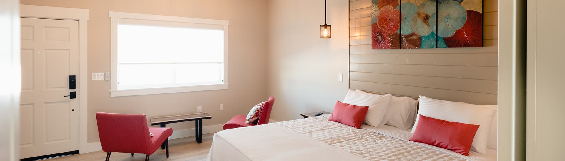 White linens on bed with fuschia floral pillows, yellow padded side chairs and dark wood table, with matching floral artwork on wall above bed.